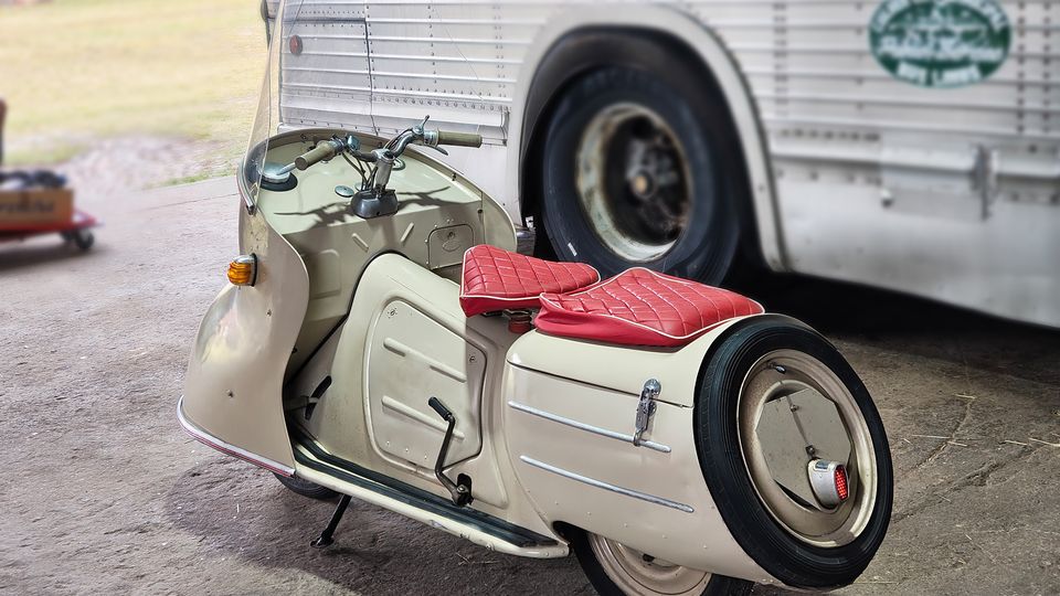 Oldtimer-Motorroller mit Seitenwagen und roten Sitzpolstern steht auf einer betonierten Fläche unter einem überdachten Bereich. Im Hintergrund ist die Seitenwand und ein großes Rad eines Busses zu sehen, dahinter eine Grünfläche.