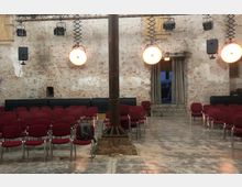 Ein Veranstaltungsraum mit Backsteinwänden, bestückt mit roten Stuhlreihen. An der Decke hängen Lampen und Lautsprecher, in der Mitte des Raumes steht eine Metallstütze, und im Hintergrund ist ein Durchgang mit Vorhängen sichtbar.