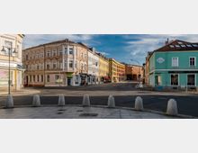 Straßenszene mit mehrstöckigen Gebäuden: Links ein historisches Gebäude mit hellroter Fassade und dekorativen Fensterrahmen, rechts ein türkisfarbenes Haus mit Ladengeschäften im Erdgeschoss. Im Hintergrund moderne, gelbe und orangefarbene Gebäude entlang einer Kreuzung.