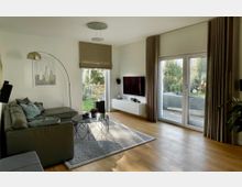 Helles, modernes Wohnzimmer mit grauem Ecksofa, Teppich und rundem Couchtisch, dazu eine Bogen-Stehlampe und ein wandmontierter Fernseher über einem weißen Lowboard. Große Glas-Terrassentüren mit Vorhängen und ein weiteres Fenster führen in einen Gartenbereich; Holzboden und neutrale Wandgestaltung prägen den Raum.