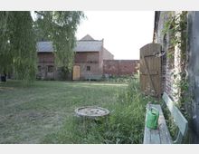 Grüne Wiese mit Bäumen und Blick auf einen Backsteinbau mit Holztüren und kleinen Fenstern; im Vordergrund eine grüne Bank, darauf eine Gießkanne, sowie ein Holztisch, umgeben von hohem Gras.