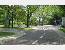 Eine kleine verkehrsberuhigte Straße inmitten eines Parks mit grünen Bäumen, Gehwegen aus Kopfsteinpflaster und Verkehrsschildern wie Zebrastreifen- und Vorfahrtsschilder. Im Hintergrund sind Sitzbänke, ein Spielplatz und Personen zu sehen.
