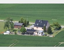 Ländliche Wohnsiedlung mit einem Einfamilienhaus in Pastellfarbe und schwarzen Dachziegeln, ergänzt durch Nebengebäude und eine kleine Gartenfläche, umgeben von weiten grünen Feldern. Im Hintergrund verläuft eine schmale Landstraße mit einigen Bäumen und Sträuchern.