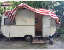 Ein kleiner, leicht abgenutzter Wohnwagen mit weißer Fassade steht auf einem gepflasterten Hof, umgeben von Bäumen und dichter Vegetation. Eine rot-weiß gestreifte Plane ist über das Dach gespannt, die Tür des Wohnwagens ist geöffnet, und davor stehen provisorische Holzstufen sowie ein Fass mit der Aufschrift „Schneider Weisse“.