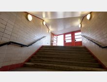 Treppenaufgang in einem Unterführungsbereich mit beige gefliesten Wänden, schwarzen Handläufen und leuchtenden runden Wandlampen. Am oberen Ende befinden sich rote Türen mit Glasfenstern, die Tageslicht hereinlassen.