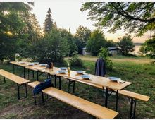 Langer Holztisch mit Sitzbänken, gedeckt mit Tellern, Gläsern und Gewürzen, steht auf einer Wiese inmitten von Bäumen. Im Hintergrund sind einzelne Häuser und ein See in einer ruhigen, bewaldeten Landschaft zu sehen.