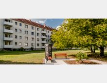 Mehrstöckiges Wohnhaus mit hellem Putz und roten Ziegeldächern im Hintergrund, daneben eine Grünfläche mit Bäumen. Im Vordergrund befinden sich ein offenes Bücherregal sowie eine Parkbank mit einem Stuhl auf einem gepflasterten Platz.