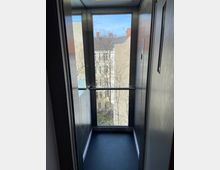 Innenraum eines Aufzugs mit Glasrückwand, die einen Ausblick auf eine Häuserfassade und Bäume mit kahlen Ästen bietet. Die Seitenwände des Aufzugs sind aus Metall, und der Boden ist dunkel.