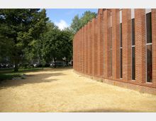 Backsteinfassade eines modernen Gebäudes mit vertikalen Fensterelementen, angrenzend an einen sandbedeckten Weg. Im Hintergrund sind Bäume und geparkte Autos entlang einer Straße zu sehen.