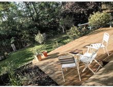 Holzterrasse mit zwei weißen Gartenstühlen und einem kleinen Tisch, umgeben von Pflanzen und Gartenmöbeln im Hintergrund. Dahinter ist eine grüne Rasenfläche mit Bäumen und Sträuchern sichtbar.