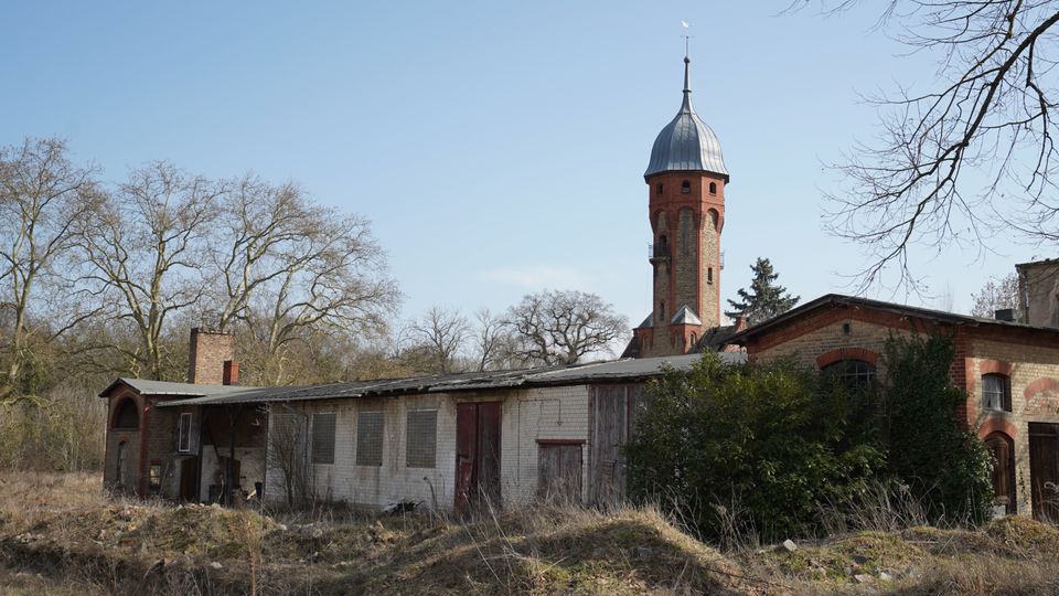 Blick auf ein Gelände mit niedrigen, teils verfallenen Backstein- und Putzgebäuden im Vordergrund; dahinter ragt ein hoher runder Backsteinturm mit Metallkuppel und Spitze auf. Umgeben ist die Anlage von trockenem Bewuchs und kahlen Bäumen unter klarem Himmel.
