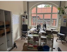 Büro mit zwei Schreibtischen, Computermonitoren, Aktenstapeln und Regalen voller Unterlagen. Ein großes Bogenfenster bietet Blick auf ein Gebäude mit Fachwerk und roten Ziegeldächern im Hintergrund.