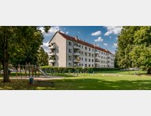 Mehrfamilienhaus mit vier Stockwerken und roten Ziegeldächern, umgeben von einer Rasenfläche mit Bäumen. Im Vordergrund befindet sich ein Kinderspielplatz mit einer Rutsche und Klettergerüst, sowie ein umzäunter Bereich mit weiteren Freizeitstrukturen.