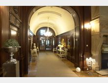 Eleganter Korridor mit gewölbter Decke, Holzwänden und dekorativen Kronleuchtern. Der Raum ist mit antiken Möbeln, Statuen und einem großen Spiegel geschmückt, und am Ende führt eine Glastür ins Freie.