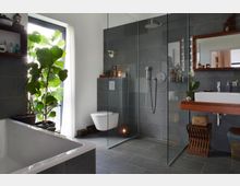 Modernes Badezimmer mit grauen Fliesen, einer ebenerdigen Glasdusche, einer freistehenden Badewanne und einem hängenden Waschbecken mit Holzablage. Ein großes Fenster mit einer Zimmerpflanze sorgt für viel Tageslicht, und dekorative Elemente wie Kerzen und Körbe ergänzen den Raum.