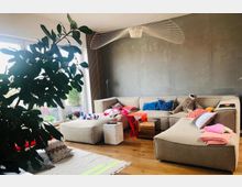 Helles Wohnzimmer mit einer großen, beigen Eckcouch, die mit bunten Kissen und Decken dekoriert ist. Im Raum stehen ein großer grüner Zimmerbaum, kleine Beistelltische und eine moderne Hängelampe, zudem ist der Boden aus Holz und eine große Fensterfront bringt viel Tageslicht herein.