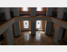 Großer Innenraum mit hohen Decken, hellorangefarbenen Wänden und grau-rosafarbenen Säulen. Drei Buntglasfenster mit abstrakten Mustern und darunter Heizkörper; im unteren Bereich stehen weiße Sitzmöbel aus Kunststoff.