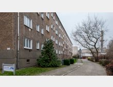 Wohngebäude mit grauer Fassade in einer Wohnsiedlung. Ein gepflasterter Gehweg führt entlang des Gebäudes, gesäumt von Büschen, einem Nadelbaum und einem kahlen Laubbaum; im Hintergrund sind industrielle Gebäude und ein Schornstein sichtbar.