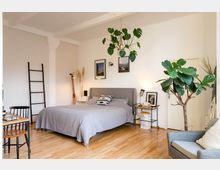 Schlafzimmer mit hellen Holzböden, einem grauen Polsterbett mit grauer Bettwäsche, einer großen hängenden Zimmerpflanze, einem Beistelltisch mit Dekoartikeln und einer Leiter an der Wand. Im Raum stehen mehrere Möbelstücke, darunter ein Sessel und ein Schreibtisch, sowie dekorative Elemente wie Wandbilder und eine Stehlampe.