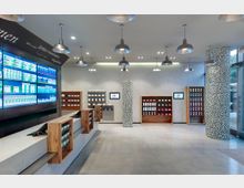 Moderner Innenraum einer Apotheke mit hellem Boden und minimalistischer Einrichtung. Zu sehen sind eine Theke mit Holzelementen, Regale mit Produkten an den Wänden und digitale Bildschirme, die Informationen anzeigen.