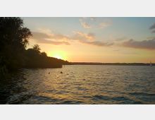 Sonnenuntergang über einem See mit ruhigem Wasser, im Vordergrund Badende nahe einem von Bäumen gesäumten Ufer, im Hintergrund ein Segelboot und eine flache Uferlinie.