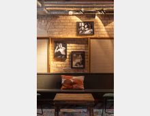 Industrie-inspiriertes Interieur mit einer Backsteinwand, an der drei schwarz-weiße Fotografien hängen. Im Vordergrund eine schwarze Holzbank mit einem orange-gemusterten Kissen, ein rustikaler Holztisch und ein farbiger Teppich auf dem Boden.