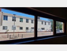 Blick durch ein Fenster auf einen zweistöckigen, weißen Putzbau mit zahlreichen Fenstern und teils abblätternder Fassade, verziert mit Graffiti. Vor dem Gebäude stehen Laternen, Poller und geparkte Autos, umgeben von gepflastertem Boden und Bäumen im Hintergrund.