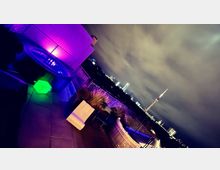 Dachterrasse bei Nacht mit beleuchtetem Whirlpool und dekorativen Leuchtelementen in Grün und Lila. Im Hintergrund ist eine Skyline mit beleuchteten Gebäuden und einem Fernsehturm zu sehen, unter einem bewölkten Himmel.