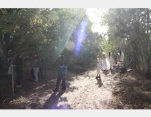 Eine bewaldete Lichtung mit mehreren Personen, darunter zwei in weißer Kleidung und andere, die sich offenbar mit Film- oder Fotoequipment beschäftigen. Sonnenstrahlen fallen durch das dichte Blätterdach, wodurch Lichtreflexionen entstehen.