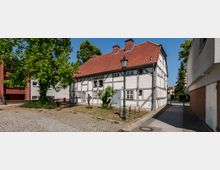 Fachwerkhaus mit rotem Ziegeldach in einer gepflasterten Hofanlage, flankiert von Bäumen und modernen Gebäuden; im Vordergrund eine Laterne und eine Bronzestatue.