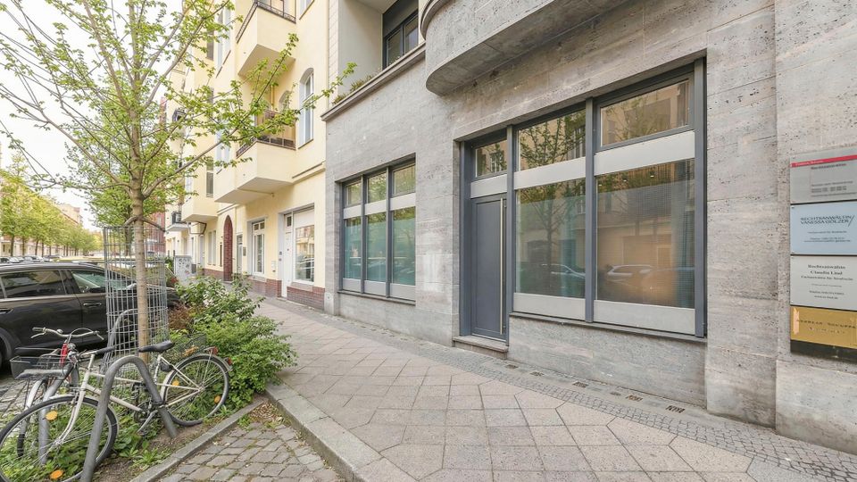 Straßenseite eines mehrstöckigen Wohn- und Geschäftshauses mit grauer Steinfassade, großen Schaufensterfenstern und einem Eingang unter einem abgerundeten Fassadenvorsprung. Vor dem Gebäude verläuft ein breiter Gehweg mit Straßenbäumen, Fahrradbügeln und geparkten Autos entlang der Allee.