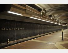 U-Bahn-Station "Haselhorst" mit dunklen Wandverkleidungen, großen metallischen Buchstaben des Stationsnamens und einer beleuchteten Decke mit markanten geometrischen Elementen. Im Vordergrund befindet sich der Bahnsteig mit Bodenfliesen in beige-braunen Farbtönen sowie ein Entwerter.
