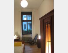 Ein einfach eingerichtetes Schlafzimmer mit hellem Holzboden, einem Bett mit gelbem Kissen, einem dunklen Schaukelstuhl und einem großen Schrank mit Holzschnitzereien. Ein Fenster mit weißen Vorhängen zeigt ein Wohngebäude im Hintergrund, und eine Deckenlampe spendet warmes Licht.