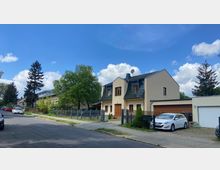 Wohngebiet mit einem beige gestrichenen Einfamilienhaus mit dunklem Schrägdach und Holzfenstern. Im Vordergrund befindet sich eine gepflasterte Auffahrt mit geparktem Auto, daneben ein Zaun und Bäume entlang der Straße unter einem blauen Himmel mit vereinzelten Wolken.