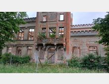 Eine verfallene historische Gebäudefassade mit freiliegendem roten Ziegelmauerwerk, zerstörten Fenstern und einem dekorativ geschnitzten Balkon über einem zugemauerten Eingangsbereich. Im Vordergrund sind hohe Gräser und Bäume sichtbar.