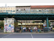 Straßenszene mit einem überdachten "Konnopke's Imbiß" unter einer grünen Hochbahntrasse in Berlin. Im Hintergrund sind mehrstöckige Altbauten zu sehen, und davor stehen Menschen in einer Warteschlange am Imbiss.