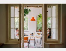 Blick durch eine geöffnete Doppeltür in einen hellen Raum mit großen Fenstern, einem weißen Tisch, Holzstühlen und hängenden Pflanzen. Eine orangefarbene Hängelampe ist zentral über dem Tisch, an dem zwei Personen sitzen.