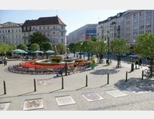 Ein kleiner Platz in einer Stadt mit einem runden Brunnen in der Mitte, umgeben von Blumenbeeten in leuchtenden Farben. Im Hintergrund sind mehrstöckige historische Gebäude, grüne Bäume, Cafés mit Sonnenschirmen und parkende Fahrräder zu sehen.