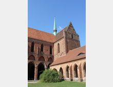Backsteinbau einer mittelalterlichen Kirche mit gotischen Spitzbogenfenstern, einem grünen Kirchturmspitz und angrenzenden Kreuzgangbögen. Der Vordergrund zeigt eine Wiese und Büsche, während der Himmel klar und blau ist.