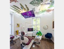 Heller Behandlungsraum einer Zahnarztpraxis mit einer modernen Behandlungseinheit, einem burgunderroten Stuhl und zahnmedizinischen Geräten. Die Decke ist mit einem bunten Dschungelmuster verziert, ein Bildschirm hängt unter der Decke, und Tageslicht strömt durch zwei große Fenster ein.