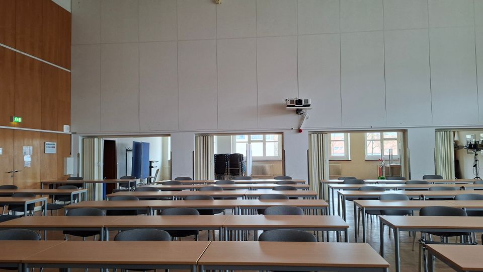 Großer Seminar- oder Konferenzraum mit mehreren Reihen langer Tische und grauen Stühlen, ausgerichtet nach vorn. Helle Wandverkleidung mit Türen zu Nebenräumen, ein an der Wand montierter Beamer sowie Fenster im Hintergrund, durch die Tageslicht einfällt.