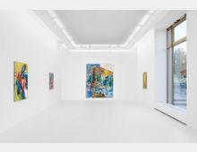 Ein moderner Galerieraum mit weißen Wänden und hellem Boden, an denen mehrere abstrakte, farbige Gemälde hängen. Große Fenster auf der rechten Seite lassen Tageslicht in den Raum.