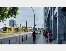 Straßenszene mit gelber Straßenbahn an einer Haltestelle, daneben Fußgänger auf einem Bürgersteig entlang eines modernen Gebäudes mit blauer Fassade. Im Hintergrund sind Oberleitungen, Ampeln und ein hohes Gebäude mit "BASF"-Logo zu sehen.