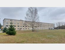 Ein vierstöckiges graues Wohn- oder Bürogebäude mit rechteckigen Fenstern, gelegen auf einer offenen Rasenfläche. Im Vordergrund stehen ein großer kahler Baum und mehrere kleine Nadelbäume, während im Hintergrund parkende Fahrzeuge und kahle Bäume erkennbar sind.