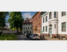 Eine ruhige Straßenansicht in einem historischen Stadtkern mit gepflastertem Weg, gesäumt von Häusern im traditionellen Stil mit verzierten Fassaden. Rechts im Bild ein weißes Gebäude mit rundem Eingang und kunstvoller Tür, links Bäume und eine niedrige Mauer mit Bögen.