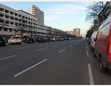 Weitläufige Stadtstraße mit mehreren Fahrspuren, links gesäumt von einem langen Wohn- und Geschäftsgebäude mit Balkonen und Geschäften im Erdgeschoss, rechts am Straßenrand geparkte Fahrzeuge. Im Hintergrund sind höhere Gebäude und Bäume sichtbar.