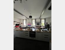 Ein geräumiger Wohnraum mit offener Küche im Vordergrund, ausgestattet mit weißen Schränken, einer Holzarbeitsplatte und einem integrierten Herd. Im Hintergrund befinden sich Ess- und Wohnbereiche mit großen Fenstern, durch die Tageslicht hereinfällt, sowie dekorative Vorhänge und verschiedene Wanddekorationen.