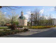 Rundkirche mit pastellrosa Fassade und grünem Kuppeldach, umgeben von blühenden Bäumen und Sträuchern in einer parkähnlichen Umgebung; im Hintergrund Wohngebäude.