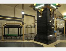 U-Bahn-Station mit Art-déco-Elementen, gekennzeichnet durch schwarz-goldene Verzierungen, glänzende Säulen und eine geometrische Gestaltung. Im Hintergrund weiße Kacheln, ein gläserner Kiosk und ein Wegweiser der Linie U7 erkennbar.