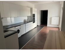 Moderne Küche mit weißen Hochglanzschränken, schwarzen Arbeitsflächen und weißer Fliesenrückwand. Der Raum verfügt über dunkle Fliesen im Küchenbereich, einen Laminatboden im Essbereich, und ist mit einem eingebauten Backofen, einer Waschmaschine sowie einem Heizkörper an der Wand ausgestattet.